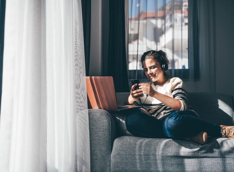 Young freelance woman works online from home in laptop sitting at comfortable sofa near balcony window behind transparent curtain