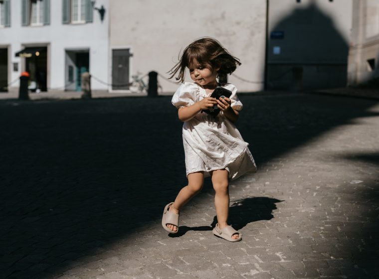 a contrast horizontal portrait of a beautiful toddler girl in motion with brown hair in a white linen summer dress holding a phone and spinning and dancing in a old town district in a spot of sunlight surrounded by shadows